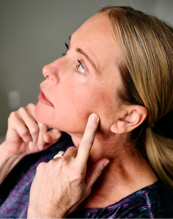 A profile shot of a blonde woman looking upward while pointing to a specific area on her cheek. She appears to be identifying a skin concern or area for treatment during a consultation. She is wearing a dark, patterned top and a silver ring. - in Tampa, FL