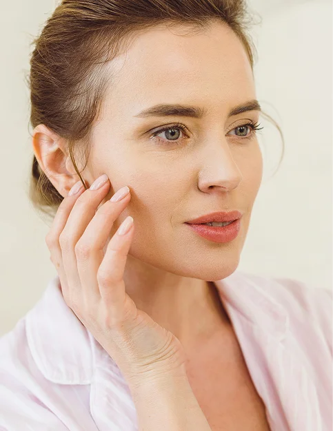 A bright, close-up lifestyle portrait of a woman with brown hair pulled back, looking slightly to the side. She has a soft smile and is touching her cheek with her fingertips, which have a neutral-colored manicure. She is wearing a light pink collared shirt. - Procedure in Tampa, FL