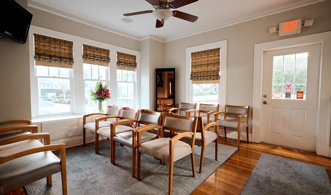 A wide view of a bright, professional waiting room featuring rows of wooden chairs with neutral cushions. Polished hardwood floors lead to a white exit door, and large windows fill the space with natural light. - in Tampa , FL