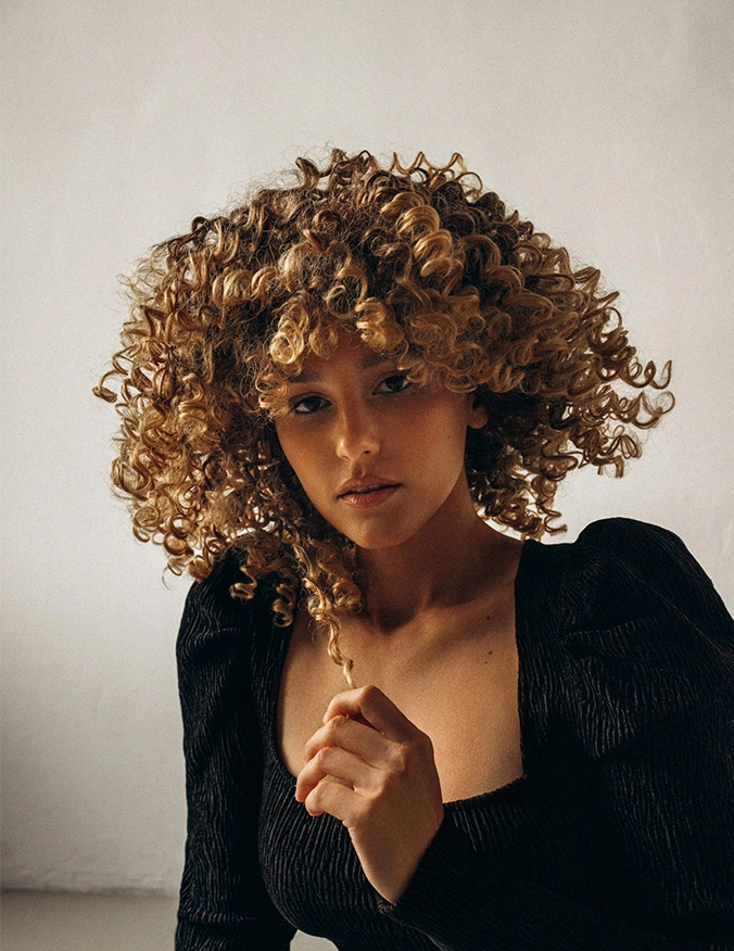 A portrait of a woman with a large, tight curly blonde afro that frames her face. She is looking forward with a neutral expression, wearing a black textured top with puffed sleeves. She holds one curl of her hair between her fingers, set against a neutral, off-white background with soft, warm lighting. - Dermal fillers in Tampa, FL