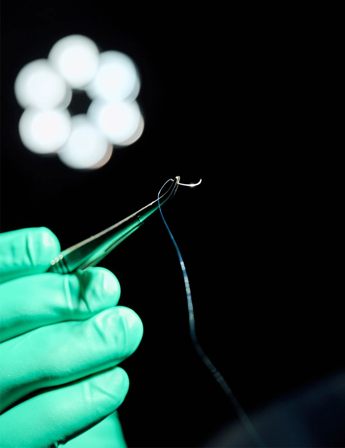 A macro photograph of a hand in a bright green surgical glove holding a pair of metal surgical tweezers. The tweezers are gripping a thin, curved surgical needle with a blue suture thread attached. In the dark background, a circular surgical light is out of focus, creating a soft bokeh effect. - Dermatochalasis and ptosis in Tampa, FL