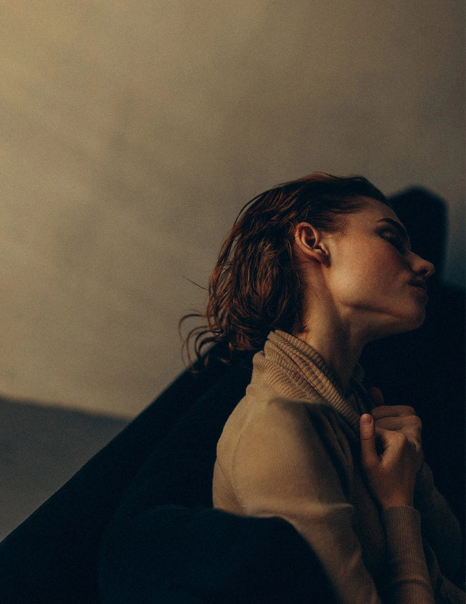 A moody, profile-view photograph of a woman with damp, reddish-brown hair swept back. She is wearing a tan turtleneck sweater and has her hands pulled up toward her neck. The lighting is low and warm, casting deep shadows that emphasize her jawline and the silhouette of her ear against a dark background. - Earlobe repair in Tampa, FL