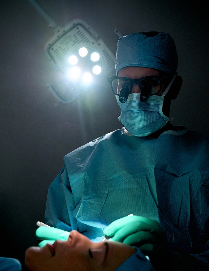 A high-contrast medical photograph of a surgeon in a darkened operating suite. The surgeon, clad in blue protective gear and a mask, looks down through magnifying loupes at a patient. Bright, focused light hits the patient's face and the surgeon’s green-gloved hands, emphasizing the precision of the clinical environment. - Ectropion surgery in Tampa, FL