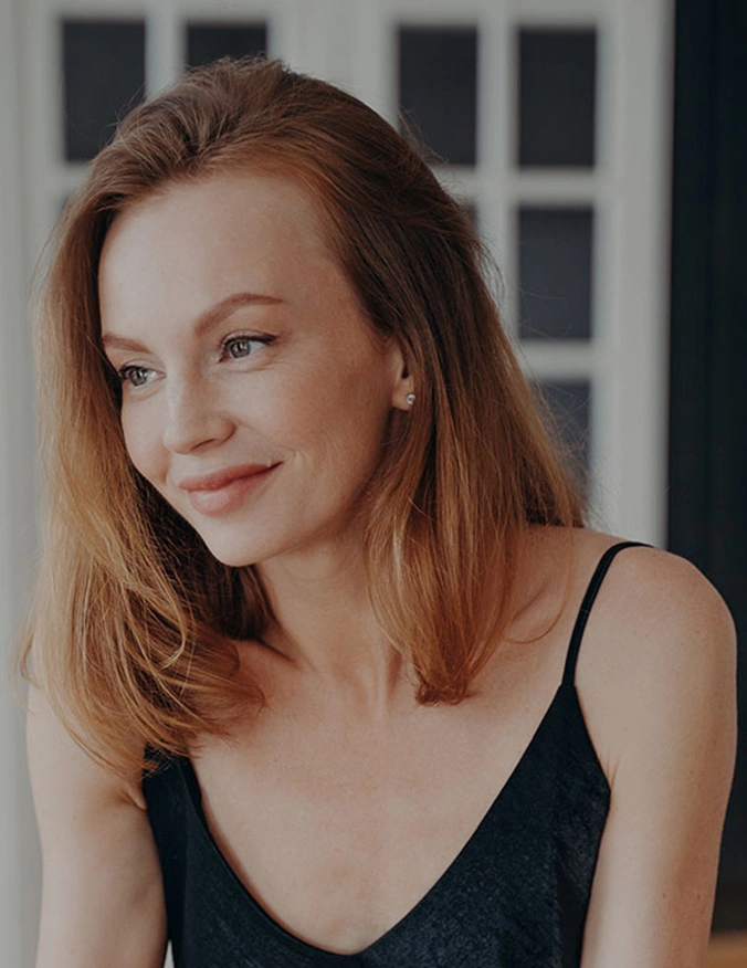 A candid-style portrait of a woman with red-toned hair and a slight smile, looking off-camera to the left. She is wearing a black spaghetti-strap top and small stud earrings. The background features a soft-focus white door with rectangular glass panes, creating a natural, lifestyle aesthetic. - Endoscopic brow lift in Tampa, FL