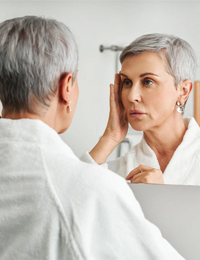 A middle-aged woman with short, stylish grey hair stands in front of a bathroom mirror wearing a white waffle-weave robe. She is looking intently at her reflection, using her fingers to gently pull back the skin at her temple to examine her eye area. The lighting is bright and clean, suggesting a morning routine. - Entropion surgery in Tampa, FL