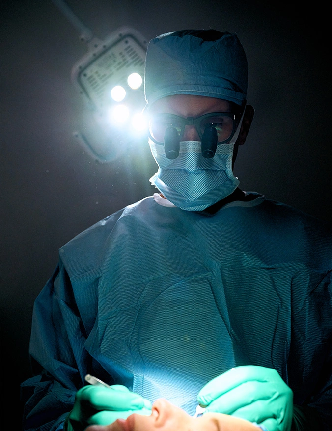 A dramatic, low-angle shot of a surgeon performing a procedure in a dark operating room. The surgeon wears blue scrubs, a surgical mask, and specialized magnifying loupes attached to their glasses. A bright surgical light overhead creates a halo effect, illuminating the surgeon's gloved hands as they work precisely on a patient’s face. - Eyelid surgery in Tampa, FL
