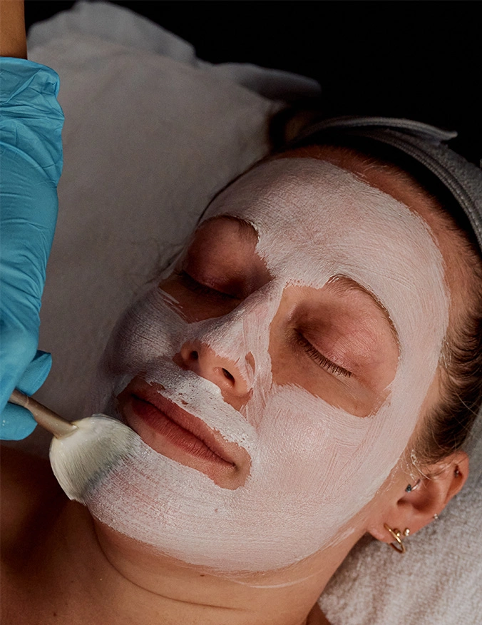 A close-up, high-angle shot of a person lying down receiving a professional facial treatment. A thick, white cream mask is being applied to their chin and jawline with a soft fan brush. A person wearing a bright blue surgical glove holds the brush, while the patient rests their head on a white towel, eyes closed in a relaxed state. - Facial peels in Tampa, FL