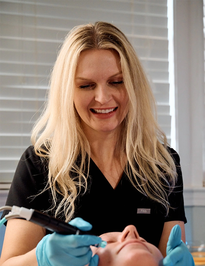 A blonde aesthetician in black "FIGS" brand scrubs smiling as she performs a facial treatment. She is wearing blue gloves and using a specialized handheld device on a client’s face. - Hydrafacial in Tampa, FL.