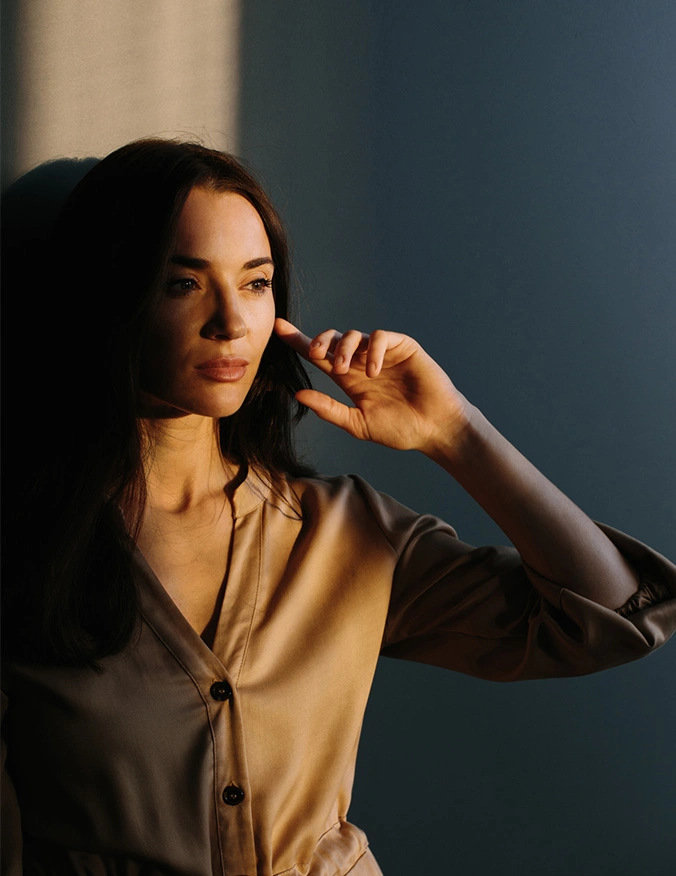 A woman with dark hair standing in soft, golden lighting that casts dramatic shadows. She is wearing a tan button-up shirt and resting a finger near her eye, highlighting smooth, clear skin. - Lasers in Tampa, FL