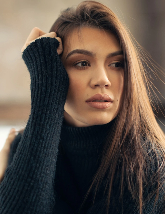 A woman with long brown hair and a soft, natural lip shape looking off-camera. She wears a dark blue ribbed turtleneck sweater and rests her head on her hand, showcasing clear skin and defined facial features. - Lip fillers in Tampa, FL
