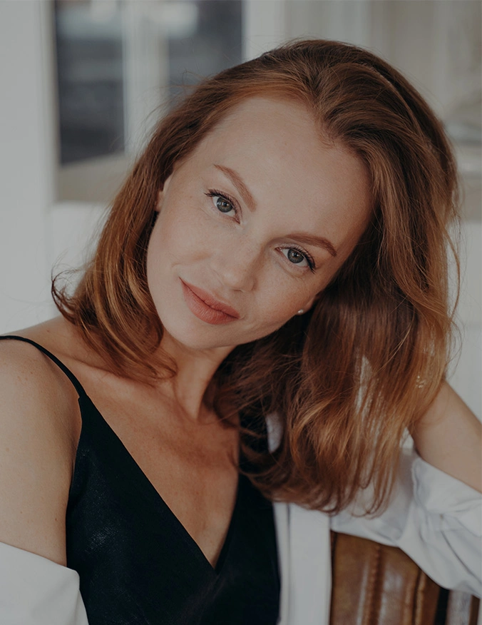 A portrait of a woman with reddish-blonde hair tilting her head slightly and smiling. She is wearing a black silk camisole and white shirt, showcasing smooth, firm skin in the lower eyelid and under-eye region. - Lower eyelid surgery in Tampa, FL