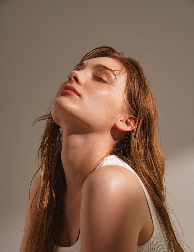 A side-profile view of a woman with long light-brown hair tilting her head back to showcase a smooth, contoured neck and a sharp, defined jawline. She is wearing a white ribbed tank top, and the warm, soft lighting emphasizes the sleek profile of her chin and neck against a neutral background. - Neck chin liposuction in Tampa, FL
