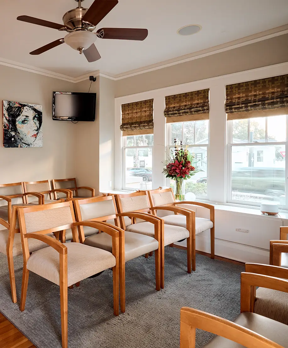 A bright and inviting medical waiting room with neutral beige walls and several rows of wooden armchairs with tan upholstery. Large windows with patterned Roman shades let in natural light, and a vase of red flowers sits on the windowsill. A flat-screen TV and a piece of modern portrait art are mounted on the wall. - In Tampa, FL