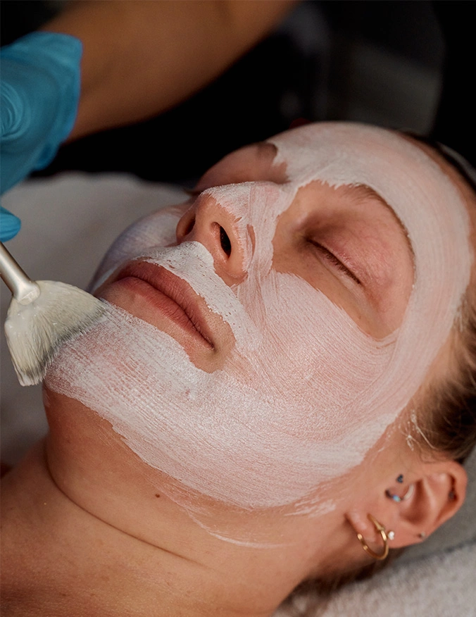A detailed close-up of a clinical skincare treatment where a white cream facial mask is being applied to a patient’s face with a professional brush. The focus is on the smooth texture of the product and the calm, relaxed state of the patient during the procedure. - Products in Tampa, FL