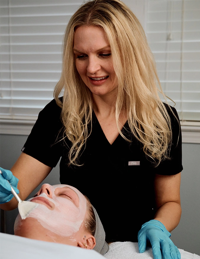 A blonde skincare professional in black scrubs and blue gloves uses a fan brush to apply a thick, white rejuvenating face mask to a client. The client is lying down with eyes closed in a relaxing clinical setting with white window blinds in the background. - Rejuvenating skin care in Tampa, FL