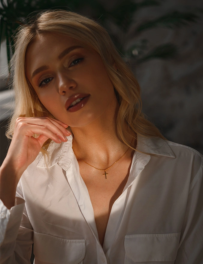 A close-up portrait of a blonde woman wearing a white button-down shirt and a small gold cross necklace. Soft, dramatic sunlight illuminates her face and neck, highlighting the firm, youthful volume of her cheeks and jawline. - Sculptra in Tampa, FL