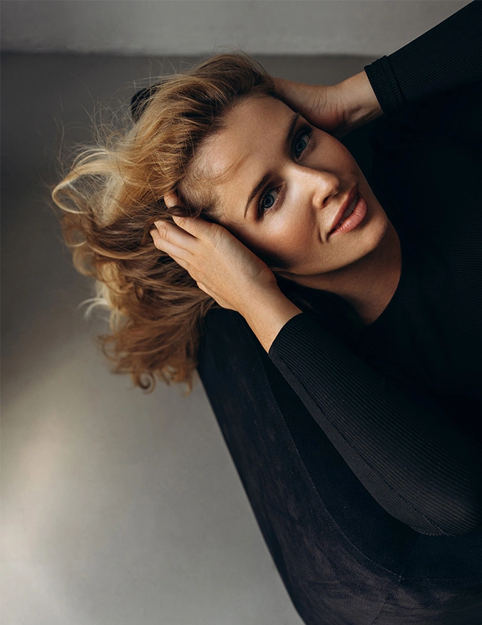 A high-angle portrait of a woman with blonde, wavy hair looking directly into the camera with clear, bright eyes. She is wearing a black ribbed sweater and has her hands gracefully tucked into her hair, emphasizing the eye area and a healthy, glowing complexion. - Tearing problems in Tampa, FL